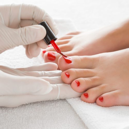 Hands in gloves cares about a woman's foot nails. Pedicure, manicure beauty salon concept. Nail varnishing in red color.